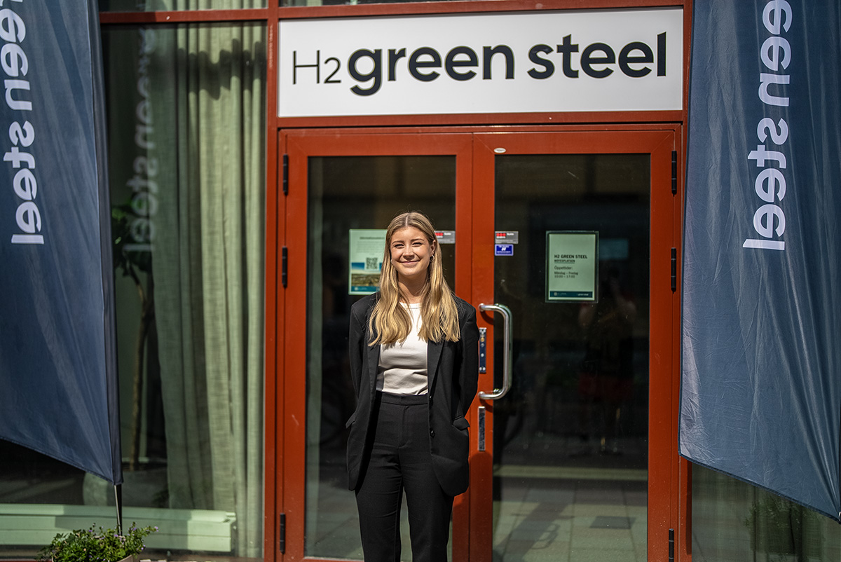 Kvinna med långt hår i svart kavaj står framför en skylt där det står H2 Green Steel